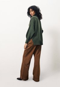 Groene gebreide trui met een hoge kraag, losse pasvorm en zijsplitten, gecombineerd met bruine broek met wijde pijpen. Soepele textuur en casual ontwerp.
