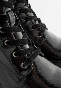 Bottes en cuir verni noir avec une texture lisse, bout rond, design à lacets et œillets en argent. Des lacets en tissu noir contrasté complètent le look.