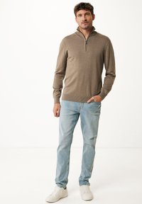 Maglione a zip marrone realizzato in tessuto morbido, abbinato a jeans azzurri e sneakers bianche. Il maglione presenta un design aderente e polsini a costine.