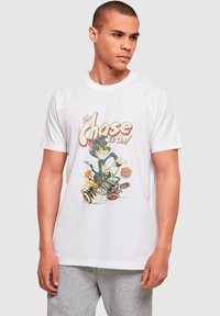 Hvid t-shirt med korte ærmer, der har et farverigt grafik af en kat og en mus i en bil, med teksten "Jagten er i gang!" ovenfor illustrationen.