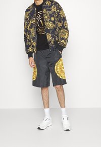 Schwarze Bomberjacke mit goldenem Barockmuster, schwarzes T-Shirt mit Logo, graue Shorts mit goldenen Akzenten und weiße Sneakers.