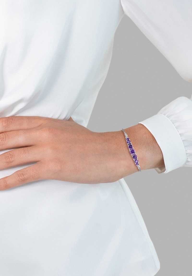 Armbånd med sølvmetalsbånd, udsmykket med jævnt fordelte lilla ædelsten. Hånd hvilende på beklædning med samlet ærmedetalje.