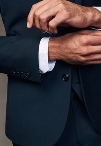 Marineblauer Anzug mit strukturiertem Finish, ausgestattet mit schwarzen Knopf-Akzenten. Weiße Hemdärmel sichtbar, teilweise hochgerollt. Hände, die den Ärmel anpassen.