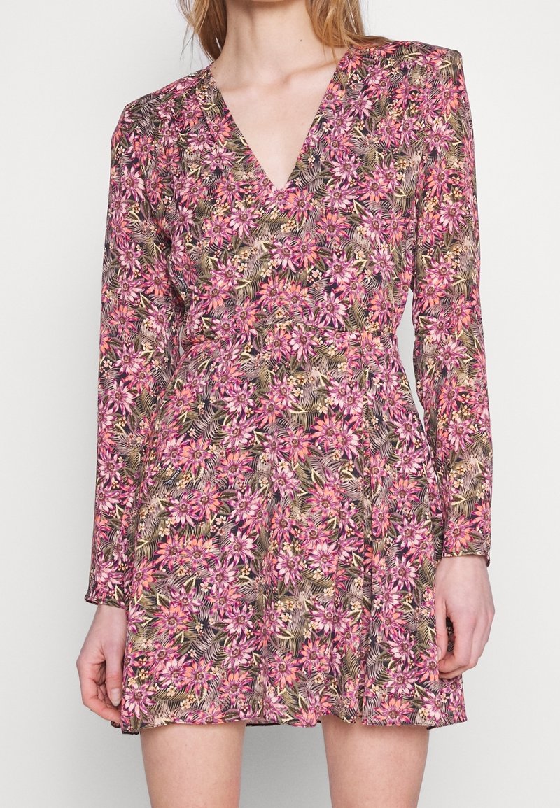 Femme portant une robe courte à manches longues et col en V, avec des motifs floraux roses et marron sur un fond clair.