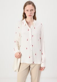 White button-up shirt with red chili pepper embroidery, long sleeves, soft fabric, and a straight cut. Paired with beige trousers and a light bag.