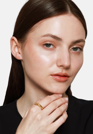 Goldene, gedrehte Ring an einer Hand, mit glatten, abgerundeten Kanten. Die Person hat blasse Haut, dezentes Make-up und helles Haar. Schwarzes Oberteil.