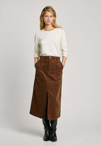 Falda midi de pana marrón con una abertura frontal, dos bolsillos y cierre de botón, combinada con una blusa clara de manga larga y botas negras hasta la rodilla.