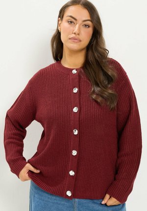 Donna con lunghi capelli castani indossa un cardigan lavorato a maglia color bordeaux con grandi bottoni argentati e jeans blu, in piedi davanti a uno sfondo chiaro e semplice.