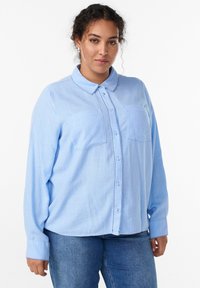 Sieviete ar cirtainiem matiem, valkājot sudraba riņķveida auskari, gaiši zilu krekla ar pogām un kabatām, kā arī zilus džinsus, stāvot pret baltu fona.