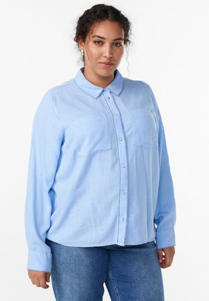 Kvinne med krøllete hår iført sølvfarvede hoops, en lyseblå skjorte med knapper og lommer, samt blå jeans, stående foran en hvid baggrund.