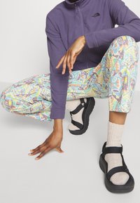 Fioletowa polarowa kurtka zapinana na zamek, wzorzyste wielokolorowe spodnie, czarne klapki ze sznurkami oraz kremowe skarpetki. Swobodny strój na świeżym powietrzu o różnych fakturach.