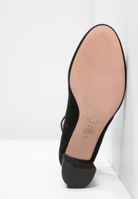 Schwarze Wildleder-Pumps mit rundem Zehenbereich, glatter Sohle und robustem Blockabsatz. Verfügt über einen Knöchelriemen und geprägtes Branding auf der Unterseite.