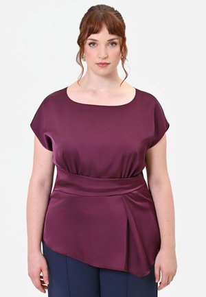 Donna dalla pelle chiara e capelli rossi indossa una blusa in raso viola scuro a maniche corte con orlo asimmetrico e dettaglio in vita alta.