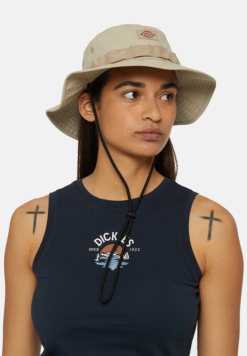 Beige Bucket Hat mit breiter Krempe, verstellbarem schwarzen Kinnriemen und strukturiertem Band um die Krone. Mit einem Logo-Patch auf der Vorderseite.