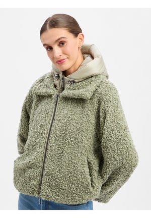 Frau mit nach hinten gegelten Haaren, die eine hellgrüne, strukturierte Fleecejacke und eine beige, gepolsterte Kapuzen-Innenschicht trägt und leicht lächelt.