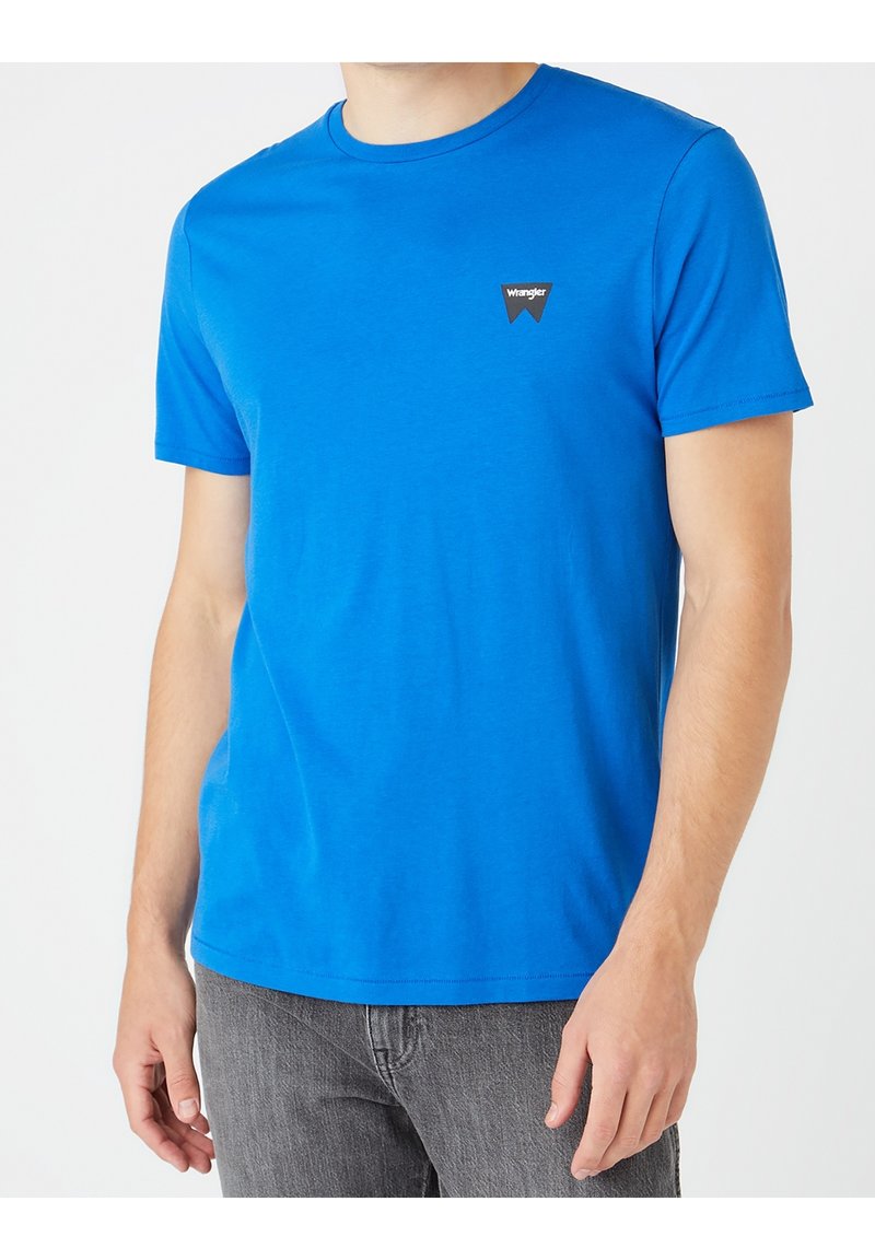 Homme portant un t-shirt à manches courtes bleu vif avec le logo Wrangler sur la poitrine et un jean gris, sur un fond neutre.