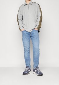 Pólder gris de manga larga con detalle de cremallera, que presenta paneles en verde oliva en los hombros. Se lleva con jeans de un azul claro y zapatillas de running oscuras.
