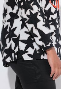 Zwart-wit top met een sterrenpatroon, lange mouwen, zijsplitsen en knoopmanchetten, gecombineerd met zwarte broek.