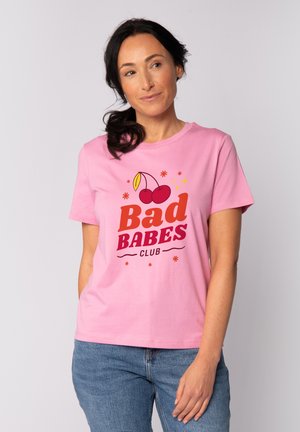 Frau mit dunklem Pferdeschwanz, die ein pinkfarbenes T-Shirt mit Kirschen und dem Schriftzug "Bad Babes Club" trägt, kombiniert mit blauen Jeans, vor einfarbigem Hintergrund.