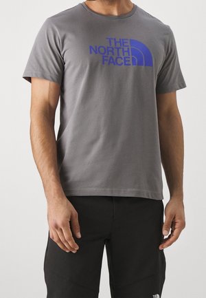 Mężczyzna w szarej koszulce z krótkim rękawem z niebieskim logo "The North Face" i czarnych spodniach stoi na jednolitym tle.