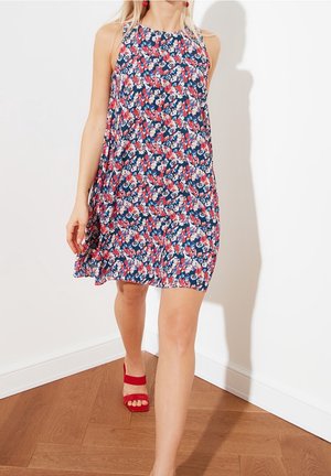 Vrouw die loopt in een mouwloze, knielange jurk met een bloemenprint in rood, wit en blauw, gecombineerd met rode open-toe hakken.