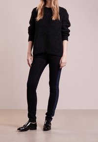 Person, der einen schwarzen grobstrickigen Pullover, schwarze Skinny-Hosen mit einem dunklen Seitenstreifen und schwarze Lederloafers trägt, auf einem glatten Boden steht.