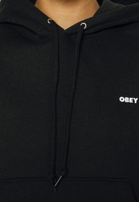 Černá bavlněná mikina s kapucí, vybavená šňůrkou, přední kapsou a bílým vyšitým logem "OBEY" na hrudi. Měkká textura s uvolněným střih.