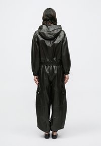 Čierny kožený overal s kapucňou, elastickým pásom a nohavicami voľného strihu. Má patentné vrecká na každej strane a rebrované manžety na členkoch.