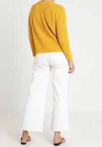 Senfgelbes Strickpullover mit langen Ärmeln, kombiniert mit weißen weiten Hosen. Hat eine entspannte Passform und eine strukturierte Verarbeitung.