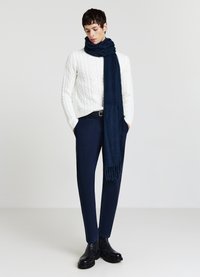 Marineblauer Wollschal, der über einem weißen Strickpullover mit Zopfmuster drapiert ist, kombiniert mit dunkler Hose und schwarzen Lederstiefeln. Schlichter Hintergrund.