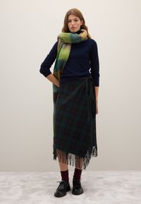 Junge Frau in marineblauem Pullover, grün-braun kariertem Wickelrock, mehrfarbigem großem Schal, burgunderroten Socken und schwarzen Schuhen steht vor einem schlichten Hintergrund.