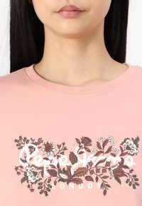 Svetloružové bavlnené tričko s okrúhlym výstrihom. Obsahuje kvetinový dizajn v hnedej a bielej farbe s textom "Pepe Jeans London" v strede.