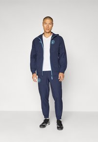 Marineblauwe windbreaker met lichtblauwe accenten en logo, gecombineerd met bijpassende joggers. De outfit heeft een witte V-hals shirt eronder.