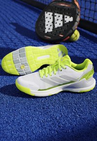 White and neon green sports shoes on blue court surface, with a black and orange paddle and yellow ball near a net in background.