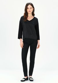 Schwarzes Oberteil mit Punkten und V-Ausschnitt, kombiniert mit schwarzen Slim-Fit-Hosen und schwarzen Schuhen mit weißen Akzenten. Glatte Texturen insgesamt.