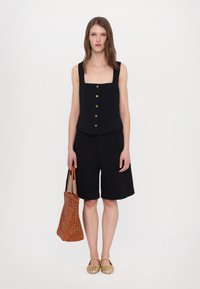 Black sleeveless vest with square neckline and gold buttons, paired with black shorts. Model holds a woven orange tote bag and wears gold slip-on shoes.
