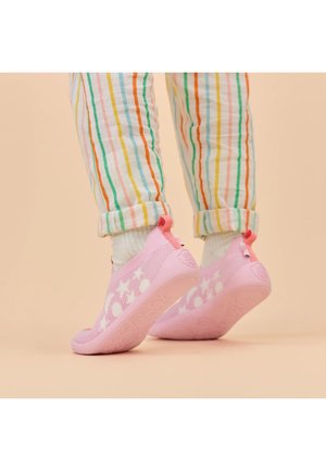 Kind draagt witte sokken, multicolor gestreepte broek en roze instappers met witte sterren- en stippenpatronen op een beige achtergrond.