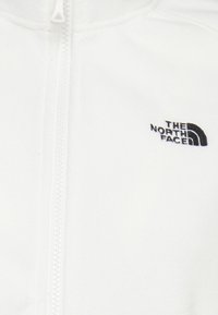 Biała kurtka z polaru z zamkiem na całej długości, z czarnym wyhaftowanym logo na lewej piersi. Miękka faktura, bez widocznych wzorów.