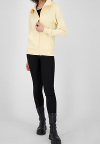 Mujer vestida con una sudadera con cremallera de color amarillo pálido, leggings negros y botas negras hasta la mitad de la pantorrilla, de pie contra un fondo liso de color gris claro.