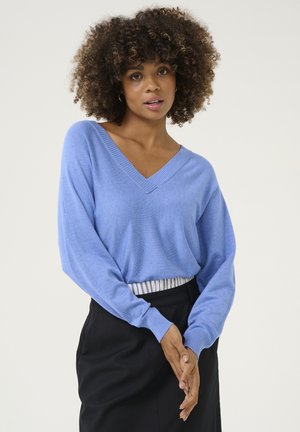 Maglione azzurro chiaro, con scollatura a V e maniche lunghe, texture morbida, indossato sopra una gonna nera con una vita a righe, che mette in risalto un fitting rilassato e una caduta fluida.