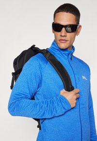 Blå jakke med glidelås laget av teksturert stoff, med høy Krage, iført en svart skulderryggsekk. Solbriller dekker øynene.