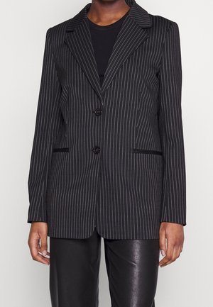 Sort blazer med nålestriber, hakkede revers, to knapper og frontlommer, båret over en sort top og sorte læderbukser.