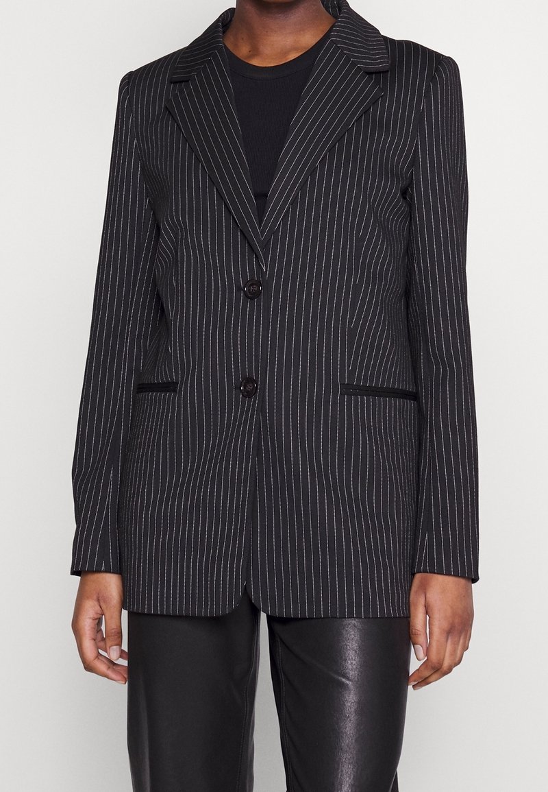 Blazer noir à fines rayures avec revers crantés, deux boutons et poches passepoilées à l'avant, porté sur un top noir et un pantalon en cuir noir.