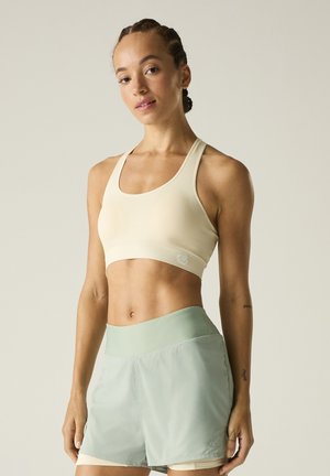 Donna con capelli intrecciati che indossa un reggiseno sportivo beige e pantaloncini sportivi verde chiaro a vita alta, in piedi davanti a uno sfondo semplice.