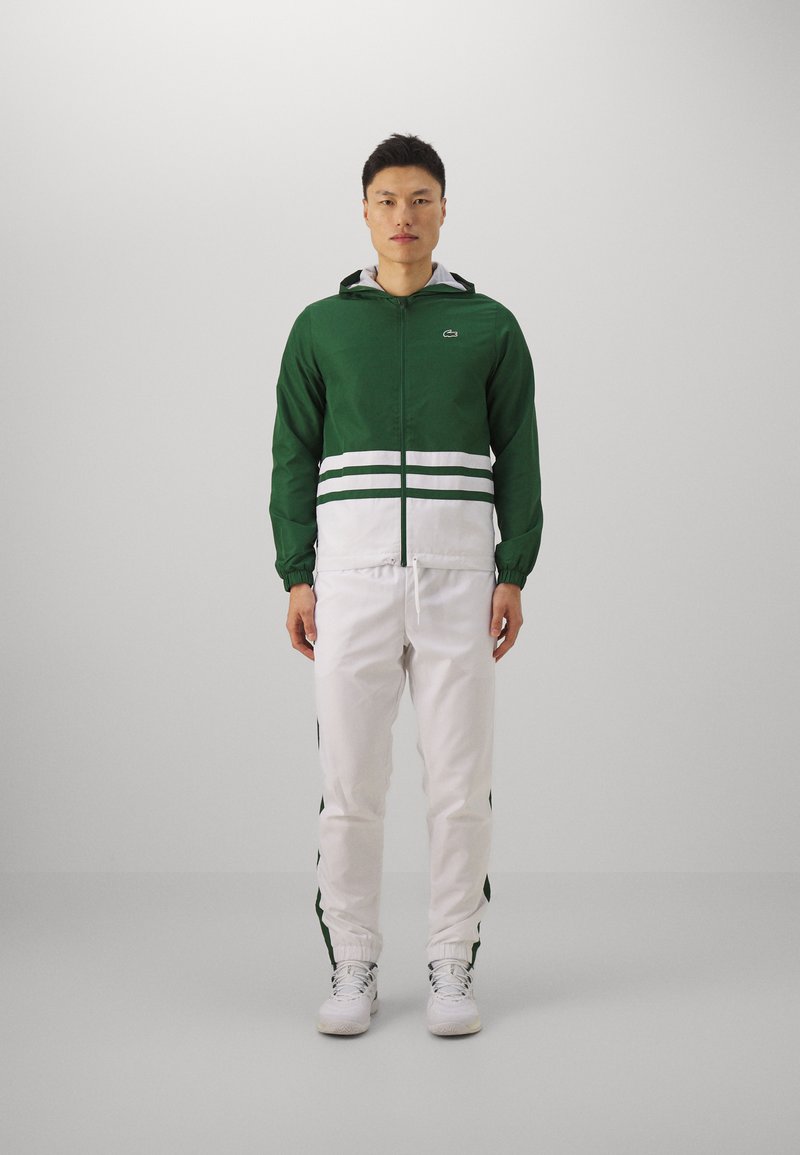 Chaqueta verde con cierre y capucha, decorada con rayas blancas, combinada con pantalones blancos que tienen acentos verdes en los laterales. El modelo se encuentra de pie frente a un fondo neutro.