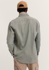 Mann, der ein hellgraues Langarmhemd, khakifarbene Cargo-Hosen und eine beigefarbene Baseballmütze trägt, mit den Händen in den Taschen stehend und von hinten betrachtet.