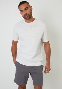 Pull en maille blanc à manches courtes, avec un motif texturé, un col rond et un ourlet contrastant, associé à un short gris.