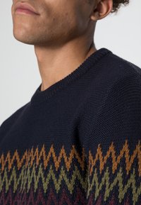 Pull en maille bleu marine avec un motif zigzag multicolore en orange brûlé, vert et bordeaux sur la moitié inférieure. Encolure ronde.