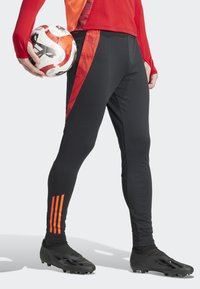 Zwarte sportbroek met een rood patroon aan de zijkant, gecombineerd met een rode lange mouwen shirt en zwarte voetbalschoenen. Er wordt een voetbal vastgehouden.