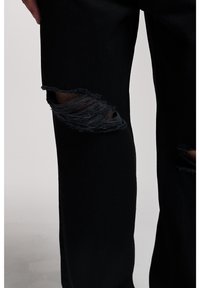 Black denim pants featuring distressed knee area with frayed edges and large rips. Material shows texture and durability.